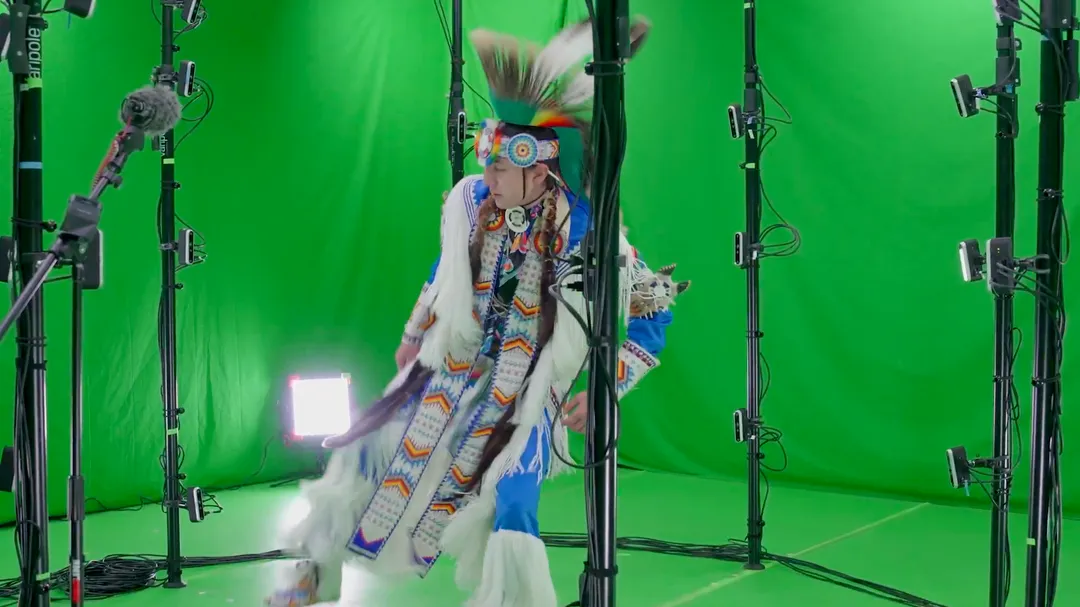 A Native American traditional dancer in full regalia performing inside a green-screen volumetric capture stage ringed with cameras and lights